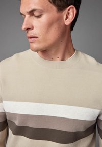 Suéter beige con rayas horizontales en blanco, marrón claro y marrón oscuro. Cuello redondo y tejido de punto texturizado. Sin logotipos visibles.