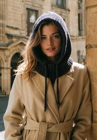 Cappotto di lana beige con vita regolabile, abbinato a un cappuccio in maglia grigio scuro con texture a costine e lacci. La luce naturale esalta i dettagli.