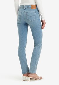 Jeans de mezclilla azul claro con corte ajustado, bolsillos traseros y una etiqueta de marca de cuero marrón. El modelo lleva sandalias blancas con tacón ancho.