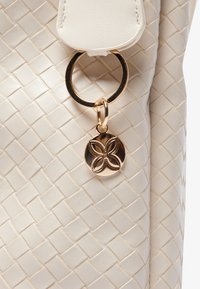 Woven faux leather handbag in off-white, featuring a gold-tone ring and a decorative gold charm with a floral design.
