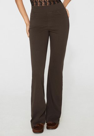 Donna con pantaloni marroni a vita alta, svasati, con tasche laterali e mocassini marroni, in piedi su uno sfondo semplice.
