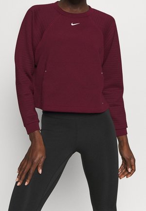 Eine Person, die ein dunkelrotes langärmliges Sportoberteil mit einem weißen Nike-Logo und schwarze enganliegende Leggings trägt, steht vor einem schlichten Hintergrund.