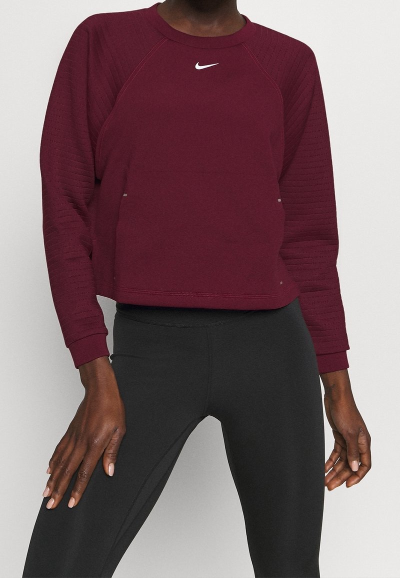 Persona che indossa una maglietta sportiva a maniche lunghe di colore rosso scuro con il logo Nike bianco e leggings neri aderenti, in piedi contro uno sfondo neutro.