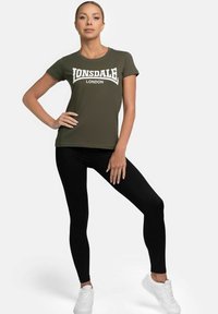 Olive green t-shirt with white "LONSDALE LONDON" text, paired with black leggings and white sneakers. Smooth fabric, fitted design.