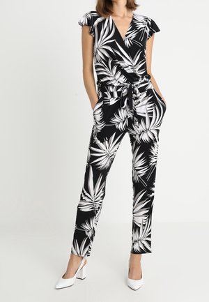 Woman wearing black and white leafy print jumpsuit with short sleeves, V-neck, tied waist, straight pants, and white heels, hands in pockets.