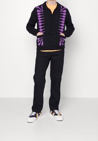 Svart kofta med lila mönster fram och fickor, bärs över en orange tröja, matchat med svarta byxor och flerskiktade sneakers.