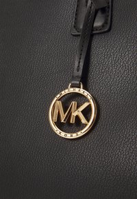 Černá strukturovaná kožená kabelka s zlatou kulatou značkou s logem „MICHAEL KORS“ a monogramem „MK“.