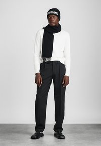 Berretto in maglia nera con logo bianco, sciarpa nera, maglione bianco, pantaloni neri su misura e mocassini neri, esposti su uno sfondo neutro.