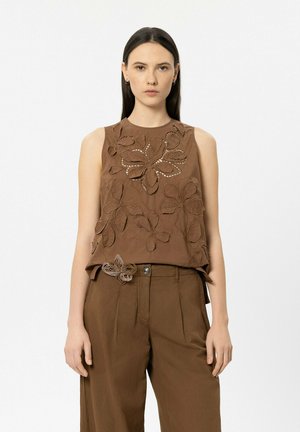 Frau trägt ärmelloses braunes Oberteil mit floralen Ausschnittmustern und passende braune Hose mit dekorativem Blumenmotiv an der Taille.