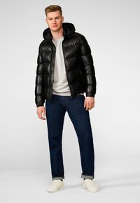 Schwarze Daunenjacke mit Kapuze, gestepptem Design und Reißverschluss vorne, getragen über einem hellgrauen Pullover, kombiniert mit blauen Jeans und weißen Sneakers.