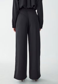 Black wide-leg trousers with a smooth, satin-like texture, featuring a high waist, structured pleats, and side pockets.