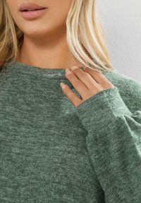 Pullover verde morbido con collo a giro e maniche lunghe. Il tessuto presenta una texture melangiata, mostrando variazioni di colore.