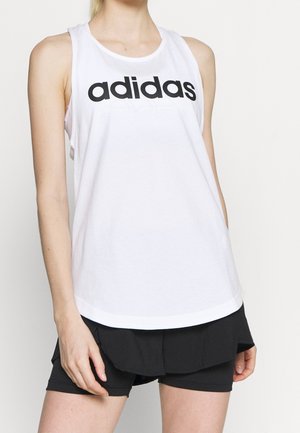 Mujer con camiseta sin mangas blanca de Adidas y pantalones cortos deportivos negros sobre un fondo claro y simple.