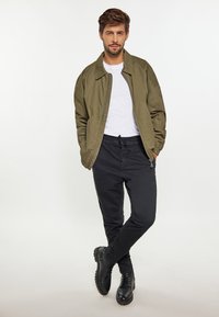 Oliven Grøn bomberjakke over en hvid t-shirt, parret med sorte slim-fit bukser og sorte snørebåndsstøvler, der har detaljer med lynlås lommer.