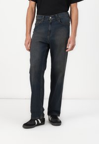 Dark blue denim jeans with a straight-leg fit, five-pocket design, and button front closure. Worn with black sneakers featuring white stripes.