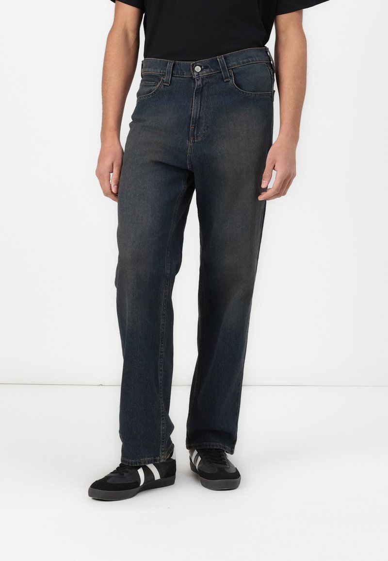 Dark blue denim jeans with a straight-leg fit, five-pocket design, and button front closure. Worn with black sneakers featuring white stripes.