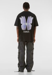 Sort oversized t-shirt med et stort blåt sommerfugl grafisk motiv på ryggen, parret med mørke cargo bukser. Teksten inkluderer "METAMORPHOSE."