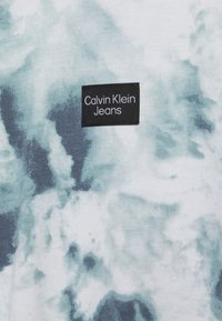 Etiqueta negra de Calvin Klein Jeans sobre tela de tie-dye en azul claro y blanco con un patrón suave, similar a nubes.