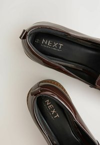 Pruuni patendi loafers, millel on ümar nina, dekoratiivne pael ja pehme sisetald. Logo lugedes "NEXT FOREVER COMFORT." Sile tekstuur.