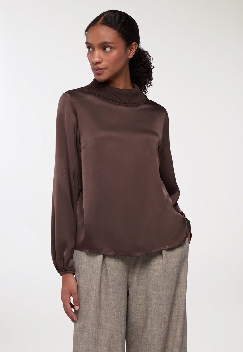 Blusa lunga in satin marrone con collo alto, caratterizzata da una texture liscia e una vestibilità rilassata, abbinata a pantaloni grigi chiari su misura.