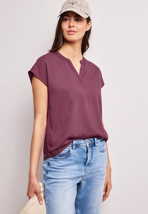 Donna che indossa un berretto beige con motivo, una blusa viola a maniche corte e jeans azzurri, che tiene in mano sinistra un tessuto di colore chiaro.