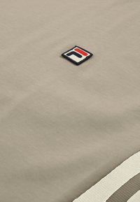 Tissu beige avec un petit patch carré présentant un logo « F » stylisé en rouge et bleu, cousu près d'un bord rayé blanc et gris.