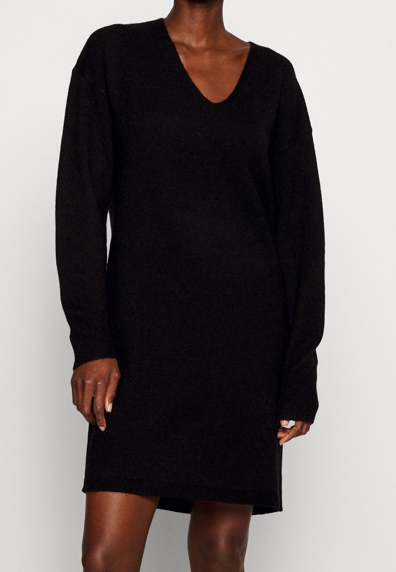 Personne portant une robe pull en maille noire à manches longues avec un col en V, debout devant un fond uni.