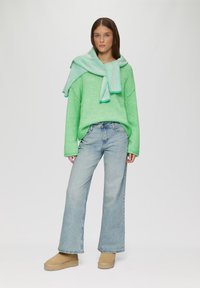 Grüner Strickpullover mit oversized Schnitt; kombiniert mit einem gestreiften, hellgrünen Schal. Verblasste, gerade Jeans und beige Stiefeletten runden den Look ab.