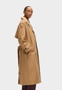 Beige Trenchcoat mit maßgeschneiderter Passform, doppelt geknöpftem Design, Schulterklappen und knöpfbaren Manschetten. Glattes Material und knielanger Schnitt.