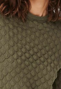 Maglione a costine verde oliva con un motivo testurizzato che presenta forme ovali in rilievo e un classico scollo rotondo. Materiale morbido e elasticizzato.