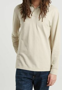 Polo beige à manches longues avec un col classique et une patte de boutonnage à trois boutons, en tissu doux. Porté avec un jean bleu foncé.