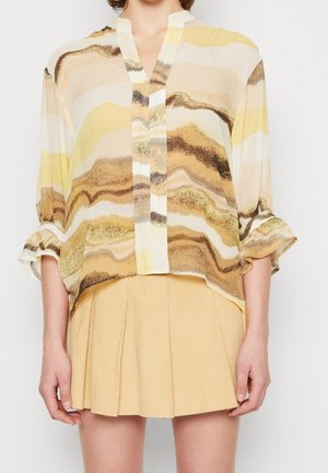 Luźna, falująca bluzka w beżowo-żółto-brązowe paski z rękawami do połowy przedramienia, zestawiona z plisowaną, jasnożółtą mini spódnicą.