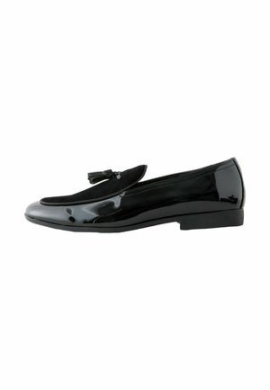 Schwarzer Lackleder-Slipper mit schwarzem Wildleder-Obermaterial und dekorativen Quasten, flacher Sohle und niedrigem Absatz, seitlich dargestellt.