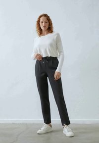 Bijela dugopruga brončana majica i tamne uske hlače s elastičnim pojasom. Model nosi bijele tenisice. Jednostavna pozadina, neutralne tonove.
