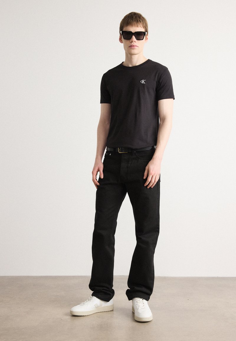 Hombre vestido con camiseta negra Calvin Klein, vaqueros negros, zapatillas blancas, cinturón negro y grandes gafas de sol oscuras, de pie frente a una pared lisa.