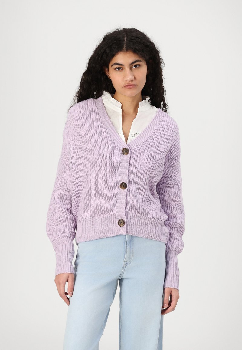 Lys lavendel strikket cardigan med dyp V-hals og tre brune knapper. Har ribbestrikkede mansjetter og en avslappet passform. Bæres over en hvit bluse.