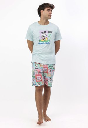 Hombre de pie descalzo, vistiendo una camiseta azul claro de Mickey Mouse y pantalones cortos coloridos con varios estampados de vacaciones temáticos de Mickey.