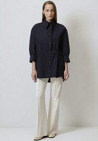 Camicia blu navy con colletto a punta e maniche arrotolate, abbinata a pantaloni crema a zampa d'elefante. Presenta un dettaglio in vita arricciato.