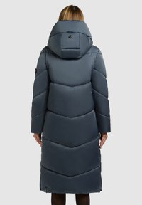 Hosszú, sötétszürke pufi kabát nagy kapucnival, halszálka mintázattal és gombos részletekkel az ujjakon. Simább, bélelt textúra.