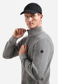 Man met een zwarte pet en een gestructureerde grijze zip-up sweater, die de rits omhoog trekt met een gefocuste uitdrukking, op een effen achtergrond.