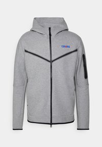 Chaqueta deportiva gris con cierre frontal, capucha y detalles en negro. Presenta un logo en azul y un bolsillo con cremallera en la manga izquierda.
