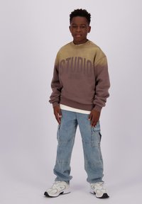 Sudadera ombre en tonos marrón y beige con el texto "STUDIO", llevada sobre una camisa blanca. Jeans cargo azules con bolsillos y zapatillas blancas.