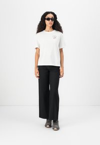 T-shirt bianco a maniche corte con una piccola grafica, abbinato a pantaloni neri a gamba larga e sneakers grigie. Texture morbide in tutto.