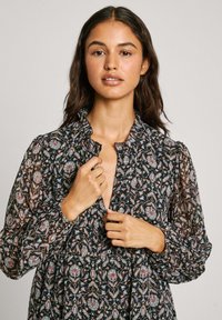 Svart blommig klänning med volangkrage, långa ärmar och knappdetaljer. Har en mönstrad design i pastellfärger på transparent tyg.