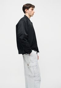 Νεαρός άνδρας με μαύρο μπουφάν bomber και ανοιχτό γκρι παντελόνι cargo στέκεται πλευρικά με το ένα χέρι στην τσέπη, πάνω σε απλό λευκό φόντο.