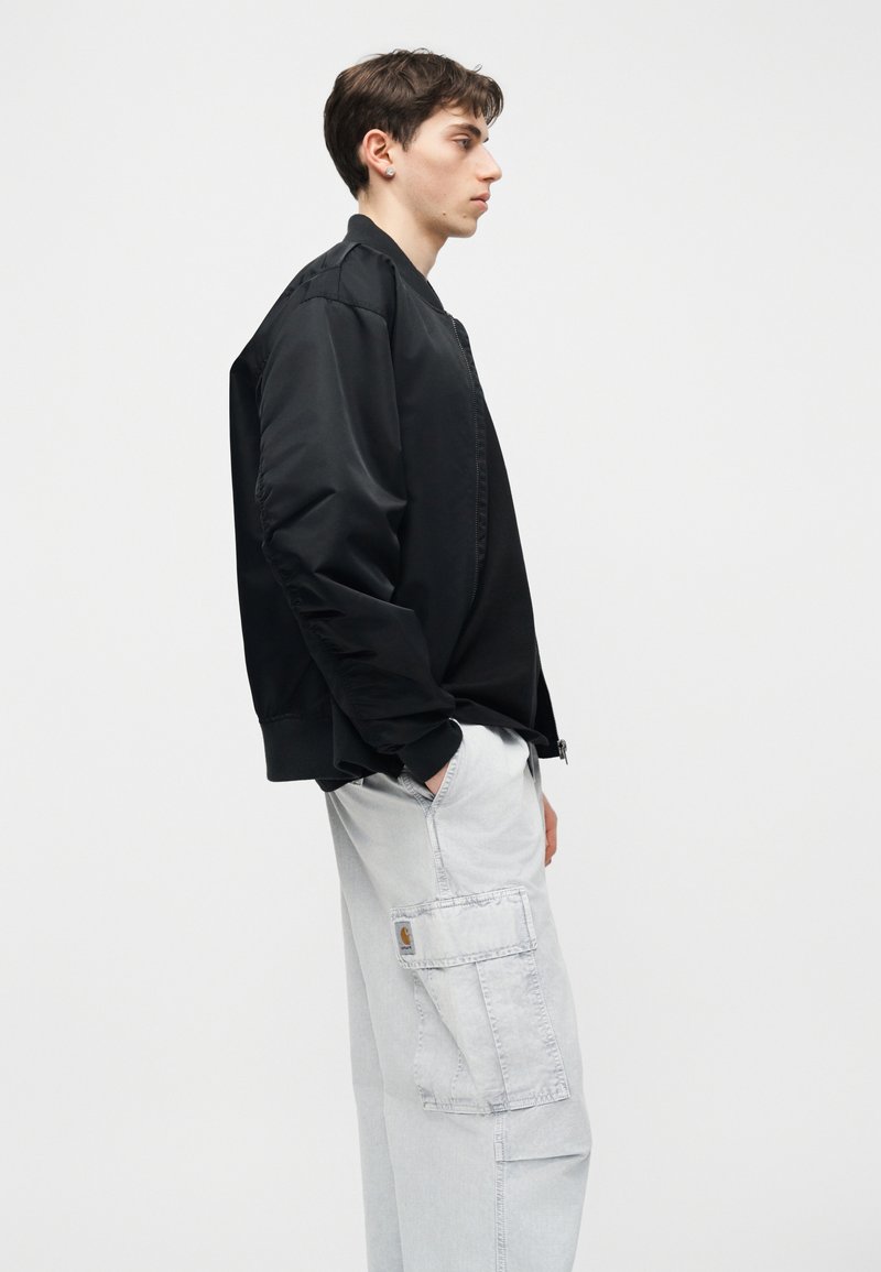 Junger Mann in schwarzer Bomberjacke und hellgrauer Cargohose steht im Profil mit einer Hand in der Tasche vor einem schlichten weißen Hintergrund.