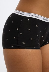 Μαύρα βαμβακερά μπόξερ σλιπ με μοτίβα από χρυσά αστέρια και λογότυπα, με λευκή ζώνη μέσης Calvin Klein. Λεία υφή με εφαρμοστό σχ έδιο.