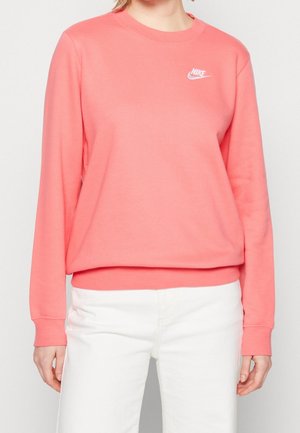 Femme portant un sweat-shirt Nike rose à manches longues et un pantalon blanc, debout devant un fond clair uni.