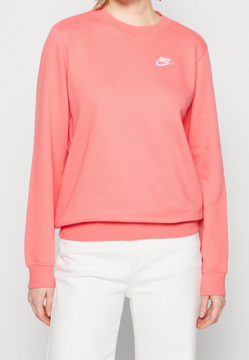 Femme portant un sweat-shirt Nike rose à manches longues et un pantalon blanc, debout devant un fond clair uni.
