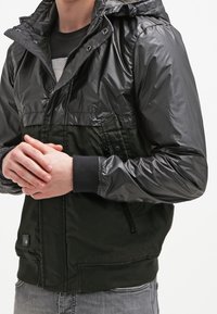 Man i en svart och mörkgrön huva-jacka med glänsande ärmar, stående med händerna knäppta framför sig.